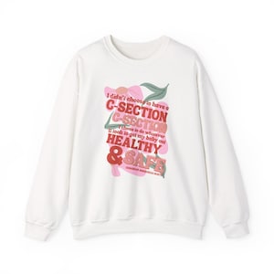 Puede incluir: Sudadera blanca con el texto "I didn't choose to have a C-SECTION" en rosa y rojo, con las palabras "HEALTHY & SAFE" en rojo y rosa. El diseño incluye un gráfico floral.