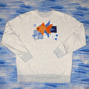 May include: A light gray crewneck sweatshirt featuring a central appliqué design of an orange goldfish layered over blue geometric shapes, including a dark blue rectangle, a blue star, and smaller blue star outlines and solid blue stars.