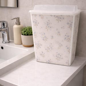 May include: A white rectangular wastebasket with a floral pattern of blue and beige flowers. The wastebasket has a clear plastic lid and is sitting on a white countertop. A sink, soap dispenser, and small potted plant are also visible.