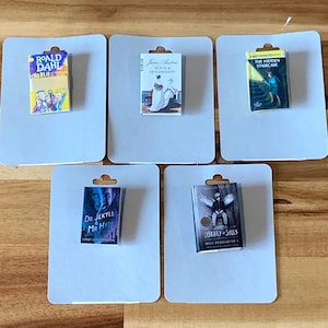 May include: Five miniature book replicas on gray cards. Titles include "Charlie" by Roald Dahl, "Sense & Sensibility" by Jane Austen, "The Hidden Staircase", "Dr. Jekyll & Mr. Hyde", and "Library of Souls". The books are displayed on a wooden surface.