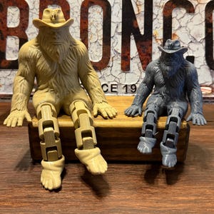 May include: Two articulated Bigfoot figurines wearing cowboy hats, one in a light tan colour and one in a dark grey colour. Both figures feature textured fur, defined facial features, and jointed legs that allow them to sit upright with feet extended.