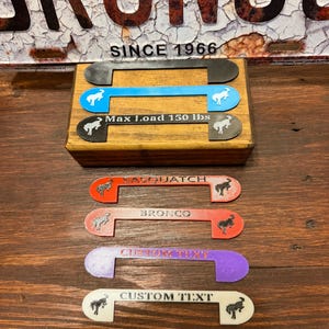 May include: A collection of U-shaped plastic handle inserts in various colors including black, blue, red, purple, and white. Several feature the Ford Bronco horse logo, while others display text such as SASQUATCH, BRONCO, CUSTOM TEXT, and Max Load 150 lbs.