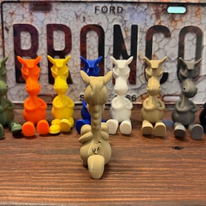 May include: A collection of eight small, stylised horse figurines in various solid colours including olive, orange, yellow, blue, white, tan, grey, and black, arranged in front of a weathered metal license plate with the text FORD BRONCO.