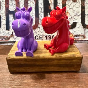May include: Two 3D printed horse figurines, one purple and one red, each featuring large eyes and a wide, toothy grin. The figures are seated upright on a rectangular, stained wooden block. The background displays a weathered metal Ford Bronco license plate.