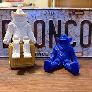 May include: Two small, articulated figures of a yeti wearing a cowboy hat. One figure is solid white and sits atop a tan square block, while the other figure is solid blue and sits directly on the surface. Both figures feature textured fur.