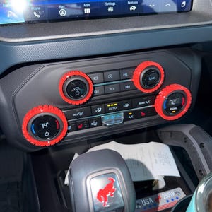 May include: A vehicle center console featuring four circular control knobs with bright red textured covers. The climate control display shows temperatures of 74 and 72 degrees. A gear shifter displays a red horse logo, and a small plaque reads USA 250TH.