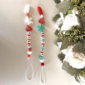 May include: Two pacifier clips featuring white, red, orange, and teal beads. One clip has white star and clownfish charms with white letter beads spelling BABY. The other has a red clip, clownfish, and teal star charms with letter beads spelling MELODY.