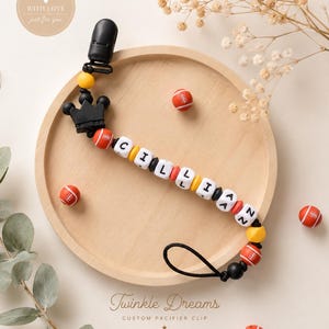 May include: A personalised pacifier clip spelling "CILLIAN" in white, black, red, and yellow beads. The clip has a black crown and football-shaped beads. The words "Twinkle Dreams" and "Handmade with Love" are also visible.