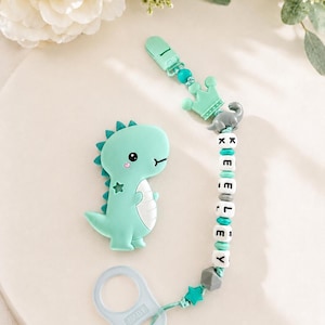 May include: A mint green baby pacifier and clip set. The pacifier is shaped like a dinosaur with a star on its side. The clip features a crown, a dinosaur, and the name "LEYLEY" spelled out with beads. The set includes a pacifier ring.