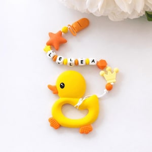 Puede incluir: Un mordedor de bebé con forma de patito de goma amarillo con el nombre "LELEA" escrito con cuentas de letras blancas. El mordedor tiene una estrella y una corona naranja, y un clip naranja. El patito tiene pies y pico naranjas.