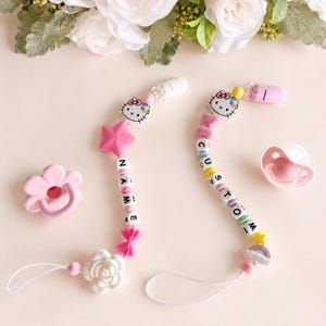 Puede incluir: Dos clips para chupetes con cuentas de silicona en tonos rosa, blanco y amarillo. Uno incluye un dije de Hello Kitty, una estrella, un lazo y una flor, con cuentas que deletrean NAME. El otro presenta un dije de Hello Kitty, un lazo y un arcoíris.