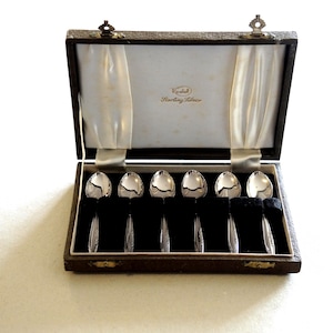 May include: A set of six silver spoons arranged in a dark brown rectangular presentation box with a white satin interior lining. The interior lid features the text "K-shell Sterling Silver" in gold lettering. The spoons have polished bowls.