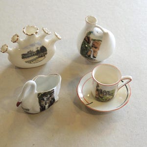 May include: A set of miniature vintage white ceramic items. Includes a multi-spouted vessel, a small jug, a swan-shaped dish, and a teacup with saucer. Each piece has a painted landscape scene and gold trim.
