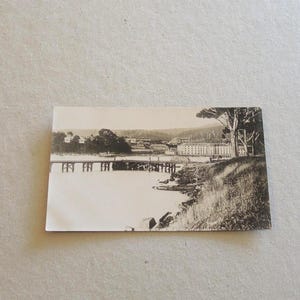 May include: A monochrome vintage photograph depicting a waterside view. A bridge spans the water, connecting to buildings on the far bank. A tree and grassy bank are seen on the right side of the image.