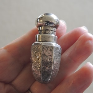 May include: A small, silver-toned metal scent bottle with a rounded cap and a cylindrical neck featuring the engraved text L&T. The body of the bottle has a tapered, acorn-like shape with vertical panels decorated with intricate, repeating floral engravings.