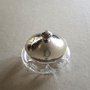 May include: A small, round glass jar with a clear, faceted star pattern on the base and a polished silver-tone metal lid. The lid features a central knob handle and is engraved with the capital letter D in a classic serif typeface.