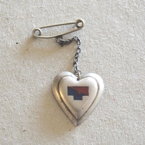 May include: A silver heart-shaped brooch with a safety pin fastening. The heart pendant has a red and blue design. A small chain links the heart to the pin. The brooch is likely vintage or antique.