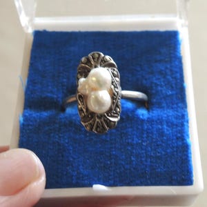 May include: A silver-toned ring featuring an elongated oval setting adorned with two white pearls and small dark grey stones. The ring is displayed in a white box with a bright blue velvet insert, showcasing a detailed metal frame around the pearls.