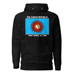 May include: Black hooded sweatshirt featuring a graphic of a conch shell within a red and blue design. The text "THE CONCH REPUBLIC" and "ANDY SANDS est. 1988" are printed above and below the graphic. The sweatshirt has a front pocket and a drawstring hood.