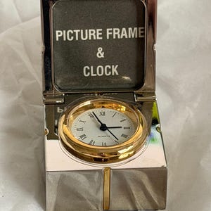 May include: A silver-toned picture frame and clock combination. The clock has a gold-colored bezel and a white face with black Roman numerals. The text "PICTURE FRAME & CLOCK" is visible on the inside of the frame.