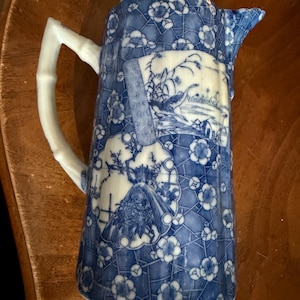 May include: Blue and white ceramic pitcher with a bamboo-style handle. The pitcher features a floral pattern and scenic illustrations. The spout is slightly chipped. A decorative piece for serving beverages or display.