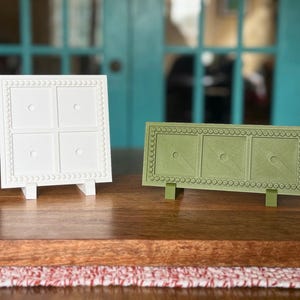May include: Two decorative trays, one white and one green, with a beaded border design. Each tray has a square or rectangular shape with a series of smaller squares or rectangles. Black handles are on either side of the trays.