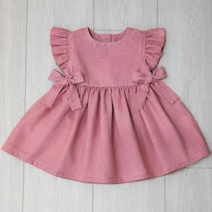 Peut inclure: Une robe rose poudré dotée d'une encolure ronde, de manches courtes à volants et d'une jupe froncée. Le vêtement est agrémenté de deux nœuds décoratifs placés à la taille de chaque côté, apportant une structure élégante au corsage.