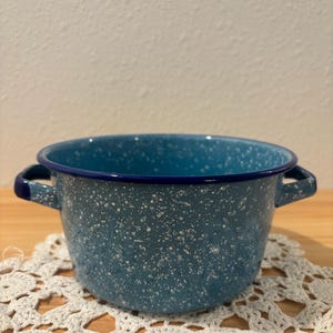 May include: A blue speckled enamelware pot with two handles. The pot has a dark blue rim and handles. The pot is sitting on a white crocheted doily. The pot is likely used for cooking.