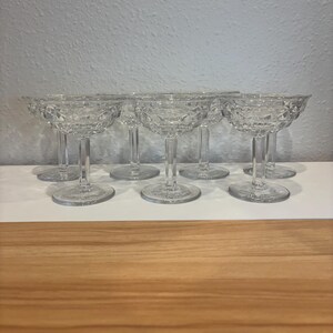 May include: A set of eight clear, stemmed champagne coupe glasses. Each glass features a round, shallow bowl with a diamond-patterned texture, supported by a slender stem and a circular base. The glasses are arranged in a row on a white surface.