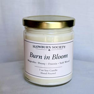 May include: A clear glass jar candle with a gold lid. The label reads "SLOWBURN SOCIETY" and "Burn in Bloom" with scent notes. The candle is 7 oz and hand poured.