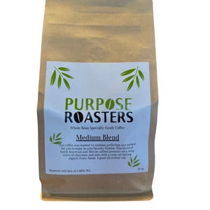 May include: A brown paper bag of whole bean coffee with a white label. The label reads "PURPOSE ROASTERS" and "Medium Blend." The text describes the coffee's flavor profile. The bag contains 12 oz of coffee.