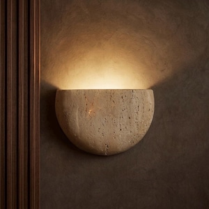 May include: A beige, semi-circular wall sconce made of textured stone. The sconce is mounted on a dark brown wall, with a warm light emanating from within. Vertical wooden panels are visible on the left side of the image.
