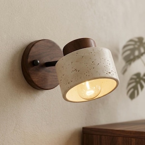 Può includere: Applique da parete moderno con un paralume cilindrico bianco sporco maculato e una lampadina calda e luminosa. L'applique presenta una base in legno scuro e un braccio nero, montati su una parete. Il design è minimalista e contemporaneo.