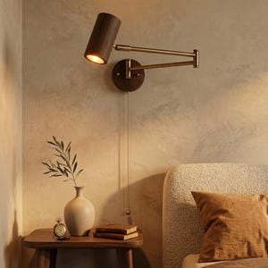 Peut inclure: Une lampe de lecture murale avec un abat-jour en bois foncé et un bras en laiton. La lampe est fixée à une base en bois ronde et possède une chaîne de traction. La lampe est positionnée au-dessus d'une petite table de chevet en bois avec un vase et des livres.