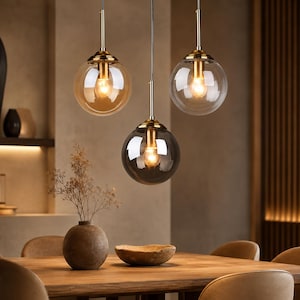 Op de afbeelding: Drie hanglampen met bolvormige glazen kappen in amber, helder en rookgrijs. Elke lamp heeft een goudkleurige metalen armatuur en een zichtbare gloeilamp. De lampen hangen boven een houten tafel en creëren een warme sfeer.