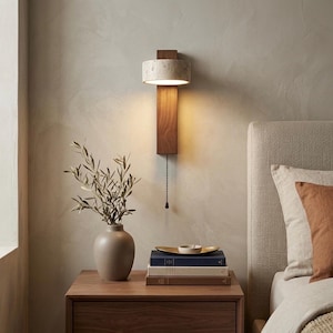 Puede incluir: Una lámpara de pared moderna con una pantalla cilíndrica moteada y una base rectangular de madera. La lámpara está montada en una pared de tonos neutros y tiene una cadena de tracción. Un jarrón con vegetación se encuentra en una mesita de noche de madera.