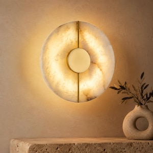 Può includere: Un'applique rotonda con una facciata in pietra traslucida color crema. L'apparecchio ha una cornice in metallo color ottone e una luce centrale circolare. È montata su una parete beige, con un vaso decorativo e una pianta in primo piano.