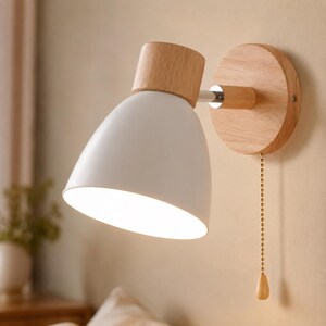 Peut inclure: Une applique murale avec un abat-jour blanc en forme de cône et une base en bois. La lampe présente un accent en bois sur le dessus et une chaînette. Le design combine des éléments modernes et naturels.