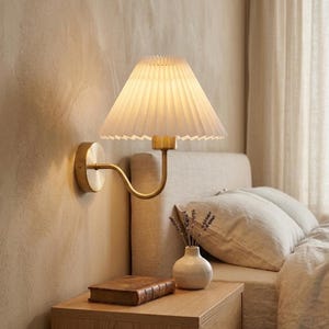 Puede incluir: Una lámpara de pared de latón con una pantalla plisada blanca. La lámpara tiene un brazo curvo y una base circular. La pantalla tiene forma de cono y emite una luz cálida. La lámpara está montada en una pared beige, ideal para un dormitorio.