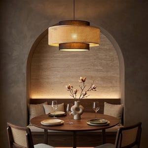 Puede incluir: Escena de comedor con una mesa redonda de madera, dos sillas y un banco empotrado. Una lámpara colgante con una pantalla de ratán tejido y una sombra oscura cuelga encima. Un jarrón con flores y copas de vino completan el ambiente.