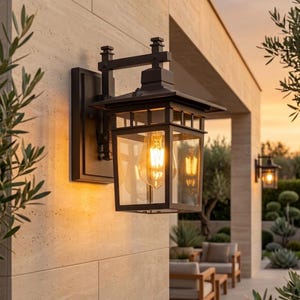 Puede incluir: Una lámpara de pared exterior negra con una carcasa de vidrio rectangular y una parte superior decorativa. La lámpara está montada en una pared de piedra de color claro. La cálida luz de la bombilla es visible a través del cristal.