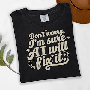Könnte beinhalten: Schwarzes T-Shirt mit cremefarbenem Text "Don't worry, I'm sure AI will fix it". Das Shirt ist gefaltet und liegt auf einer weißen Oberfläche. Ein brauner Hut ist im Hintergrund zu sehen.