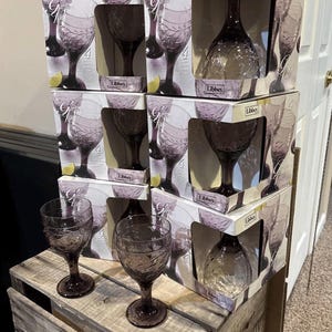 May include: A stack of Libbey Garden Vine glassware boxes containing purple-tinted wine glasses with embossed vine patterns. Two matching purple wine glasses with textured vine designs stand in front of the boxes on a wooden surface.