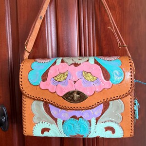 May include: A tan leather handbag featuring hand-tooled floral patterns painted in shades of pink, turquoise, and lavender. The bag has a single shoulder strap, a brass-toned turn-lock closure, and decorative whip-stitched edges along the flap.