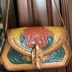 May include: A tan leather shoulder bag featuring a hand-tooled floral design on the front flap. The embossed pattern is accented with painted sections in yellow, red, and green. The bag has a braided leather edge and a central buckle strap closure.
