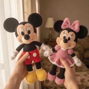 Op de afbeelding: Twee handgemaakte gehaakte poppen van klassieke tekenfilmfiguren. De ene pop heeft zwarte oren, een perzikkleurig gezicht, een zwart lijfje, een rode broek met witte knopen en gele schoenen. De andere pop heeft zwarte oren en een roze strik.
