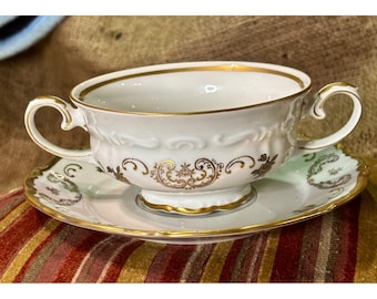 Puede incluir: Un juego de taza de caldo y plato de porcelana blanca con dos asas ornamentadas y un delicado patrón de filigrana dorada. El borde de la taza y del plato está decorado con una fina banda dorada que resalta los relieves esculpidos.