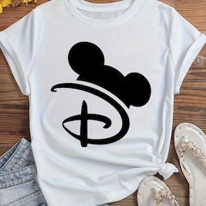 Puede incluir: Camiseta blanca con un gr&aacute;fico negro del logotipo de Disney y orejas de Mickey Mouse. La camiseta tiene mangas cortas y cuello redondo. La camiseta est&aacute; anudada a la cintura. Se ven sandalias blancas con adornos de perlas.