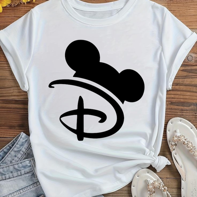 Puede incluir: Camiseta blanca con un gr&aacute;fico negro del logotipo de Disney y orejas de Mickey Mouse. La camiseta tiene mangas cortas y cuello redondo. La camiseta est&aacute; anudada a la cintura. Se ven sandalias blancas con adornos de perlas.
