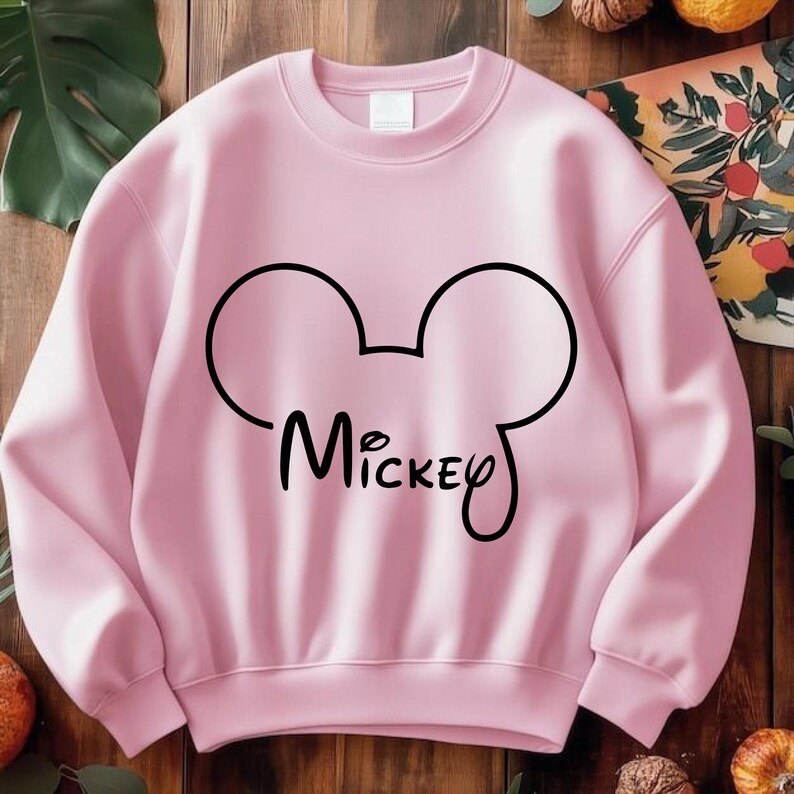 Puede incluir: Sudadera rosa claro con un contorno negro de la cabeza de Mickey Mouse y el nombre "Mickey" en cursiva. La sudadera es de cuello redondo.